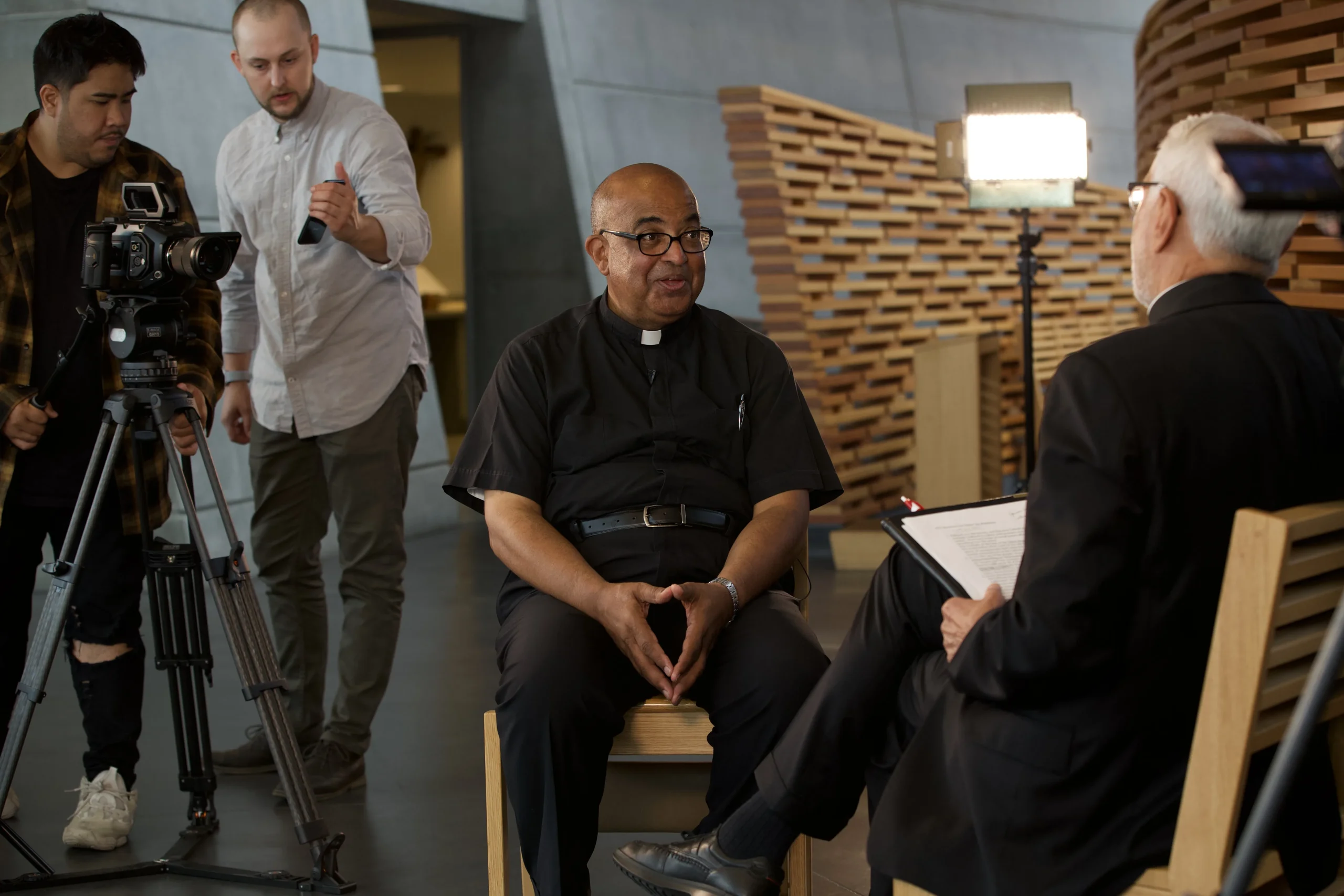 Priest interviewed in front of a wood slat backdrop